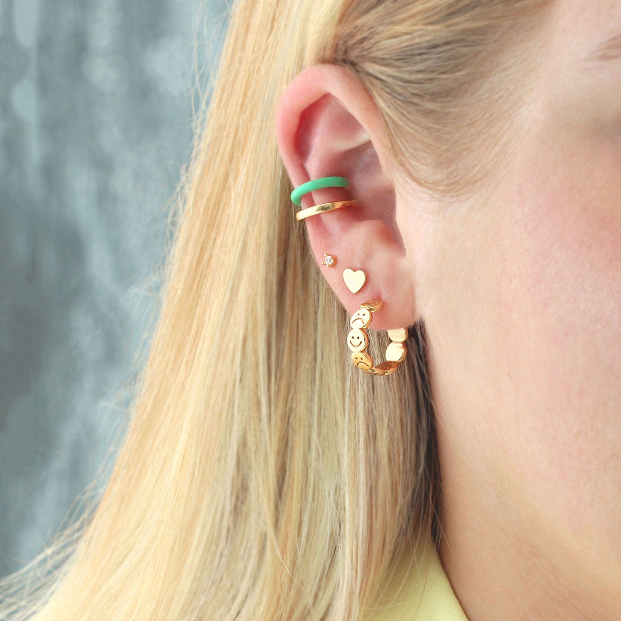 Hoop earrings styled on a model with blonde hair. The hoops are in the first piercing, with a heart stud earring in second piercing and a cubic zirconia diamond stud in the third piercing with 2 ear cuffs on top- one is gold and the other is a turquoise color made from enamel.