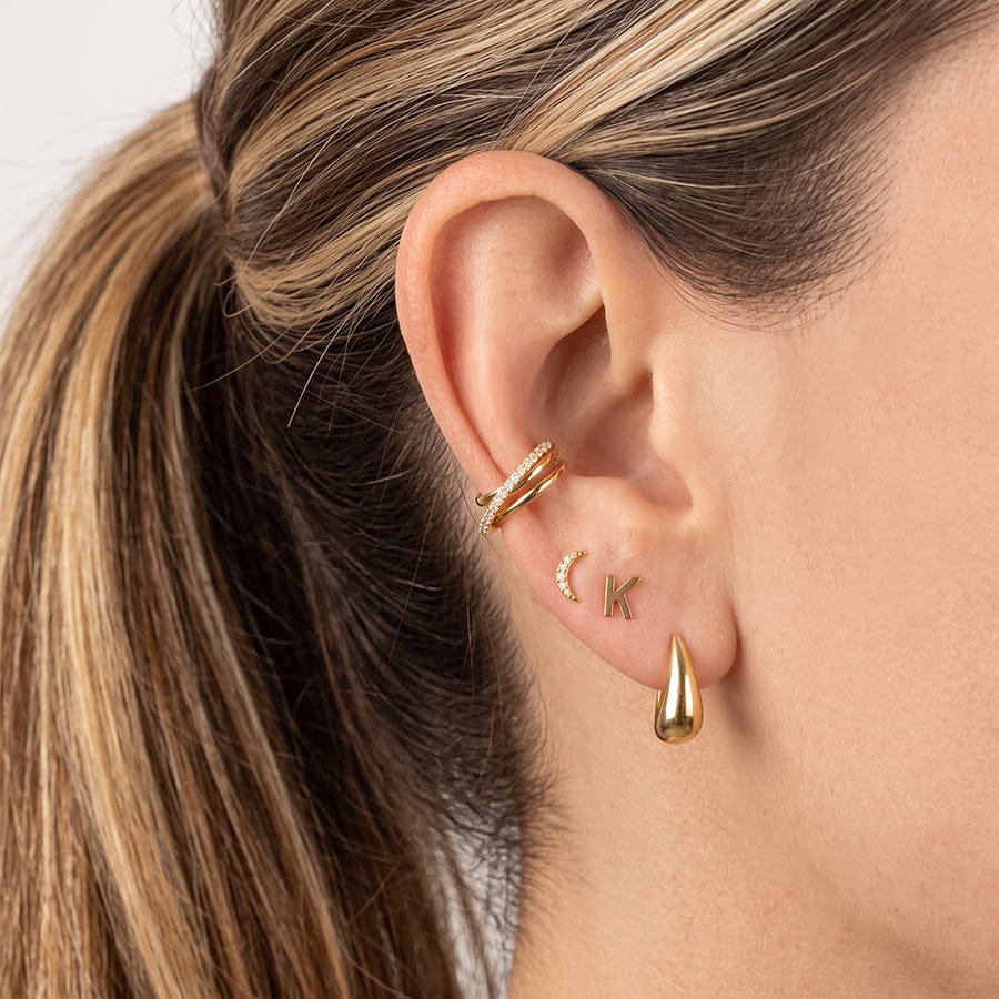 A close-up of a person's ear wearing multiple gold ear piercings: a stacked barbell hoop at the lobe, a small crescent moon stud, an initial 'K' stud, and a teardrop-shaped drop earring.
