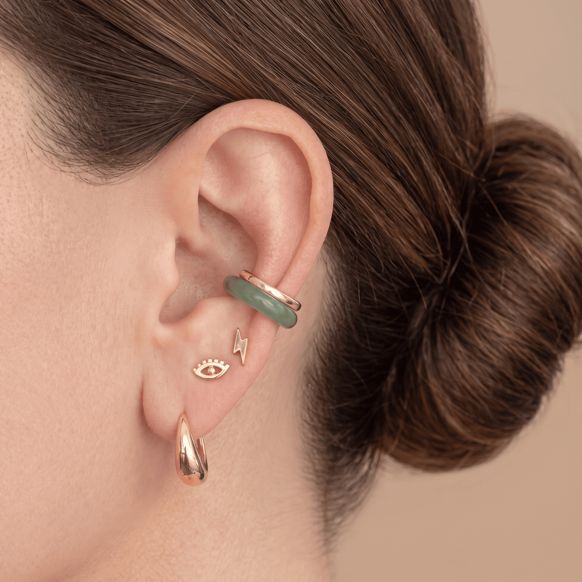 Close-up of a person's ear wearing multiple gold-toned ear piercings, including a crescent hoop, a small eye stud, a lightning bolt stud, and a green stone ear cuff, against a warm beige background.