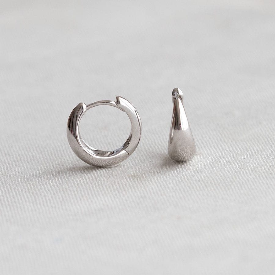 A pair of polished silver-toned earrings on a light gray textured fabric background. One earring is a small hoop, and the other is a teardrop-shaped stud.