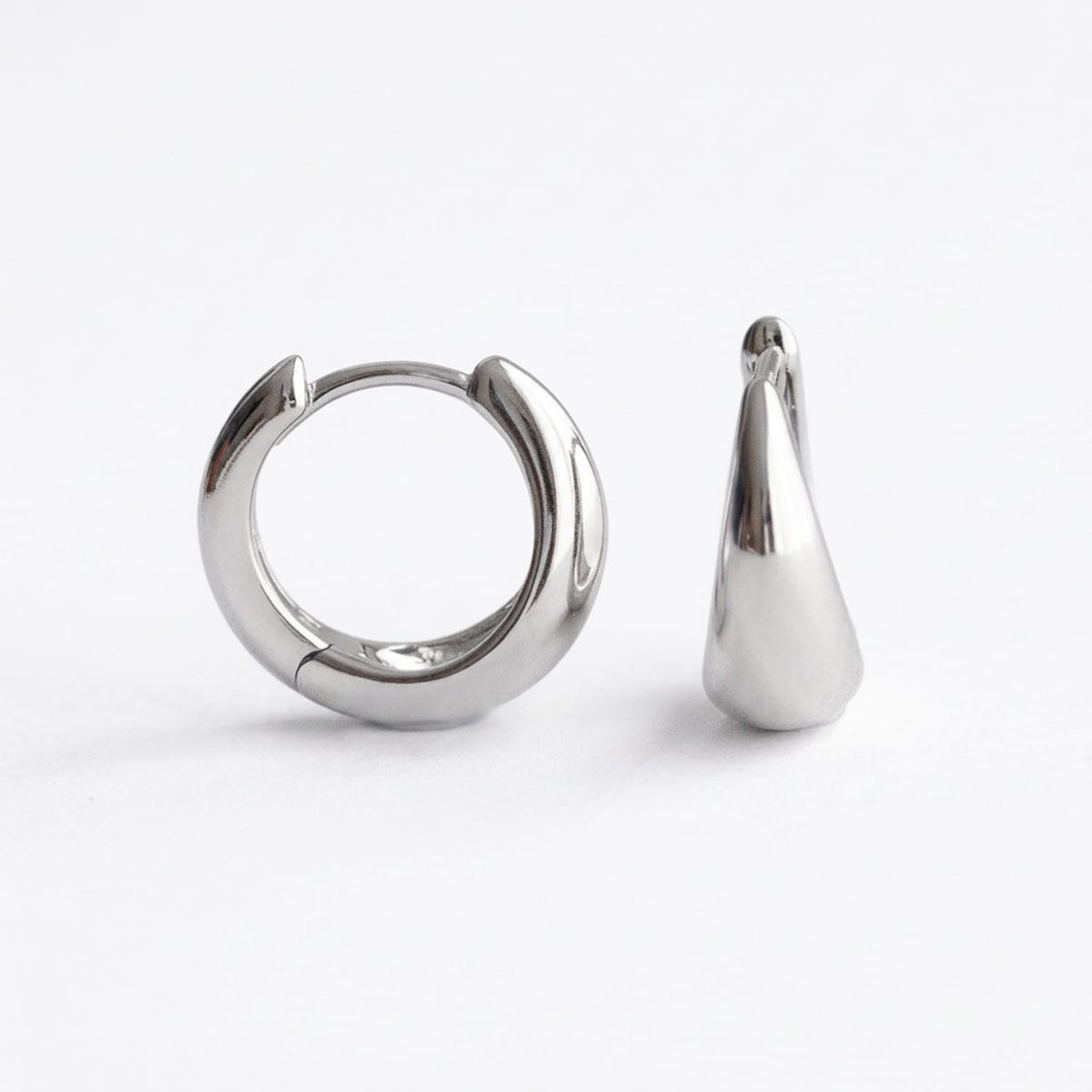 A pair of polished silver-toned hoop earrings, one shown from the front and the other from the side profile, on a clean white background.